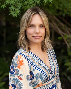 Woman with light brown hair in a floral patterned dress posing in front of trees/shrubs