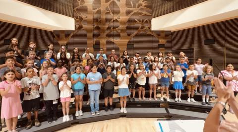 A group of school children standing on choir risers singing on a large stage.
